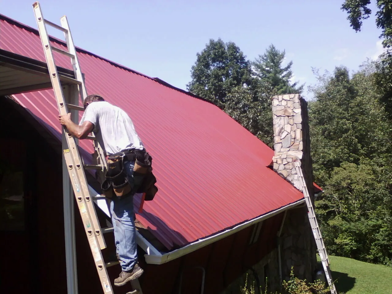 Roofer on a ladder beside a red metal roof and stone chimney — Soffit & Fascia Repair by Roofing Brothers in Dawson Springs
